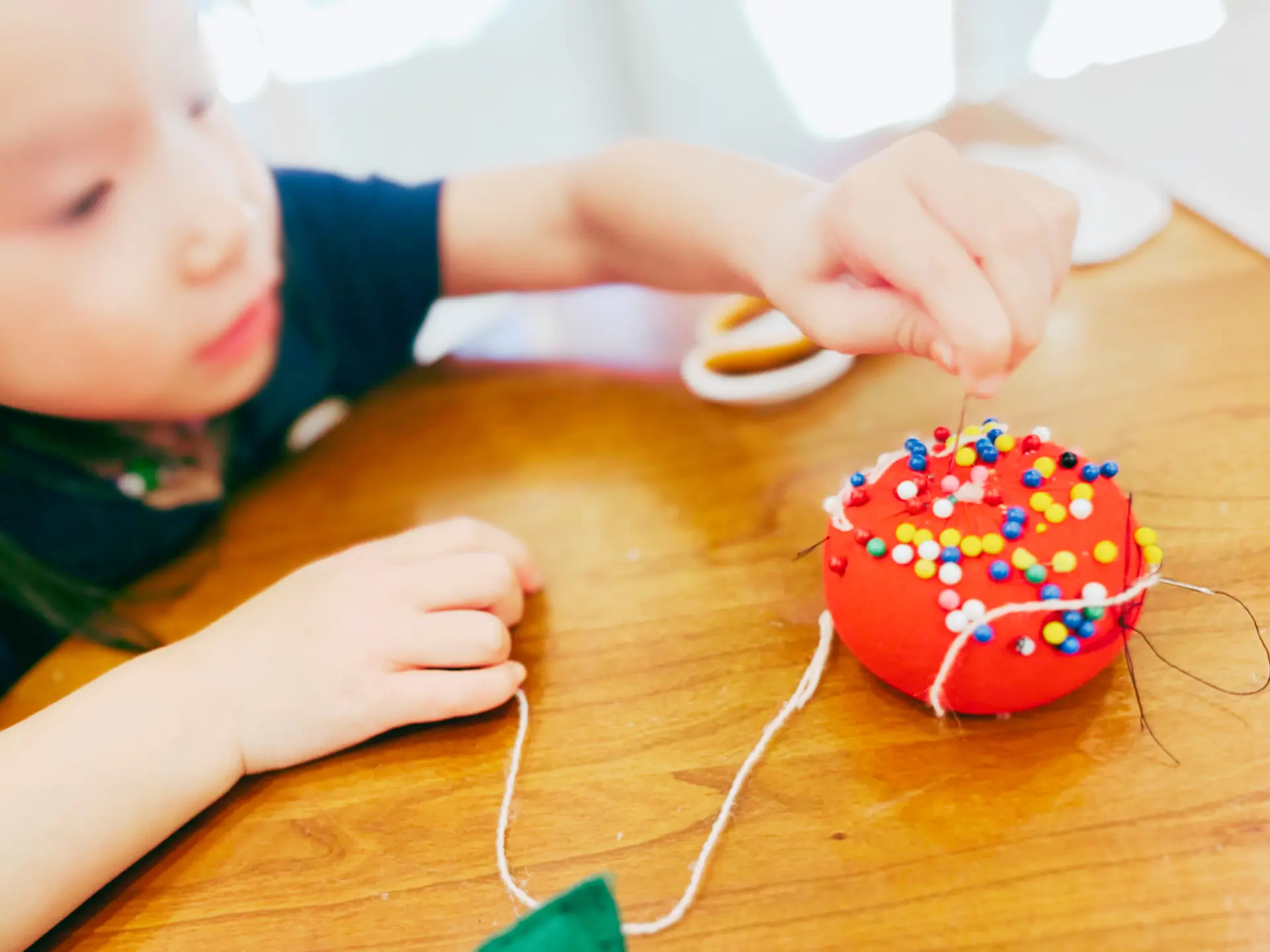 A young child carefully stitching thread through a red tomato pincushion at a wooden table